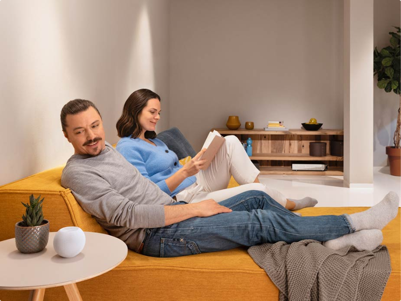Ein Paar sitzt auf dem Sofa, sie ließt und er erteilt einen Sprachbefehl an einen Sprachassistenten.