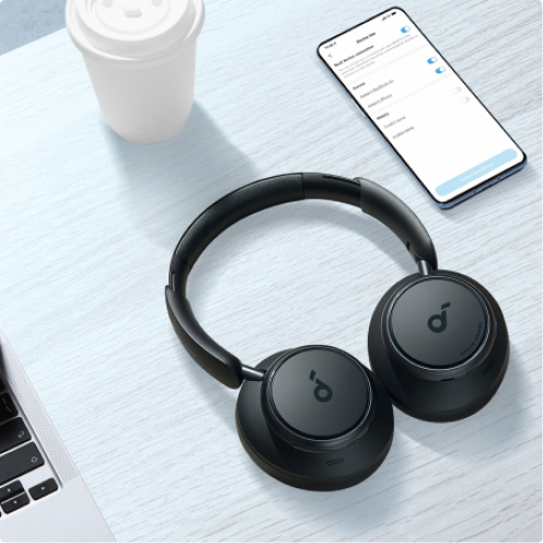 Die Soundcore Space Q45 Kopfhörer liegen auf dem Schreibtisch neben einem Smartphone, einem Laptop und einem Kaffeebecher. 