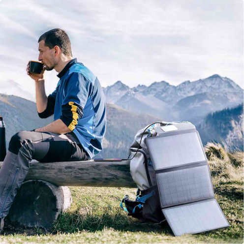 Ein Mann läd während einer Pause seiner Wanderung sein Mobilgerät auf mithilfe eines mobilen Solarmoduls von Anker, welches an seinem Rucksack befestigt ist.