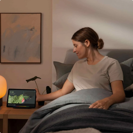 Frau im Bett stellt Google Pixel Tablet aufs Ladedock auf dem Nachttisch