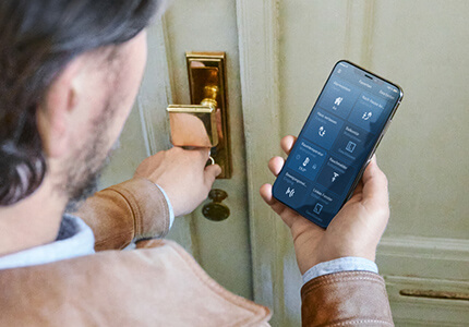 Mann steht vor Haustür mit gezücktem Schlüssel und checkt über Smartphone das Smart Home.