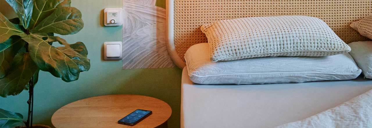 Wandthermostat im Schlafzimmer über dem Nachttisch, auf diesem liegt ein Smartphone.